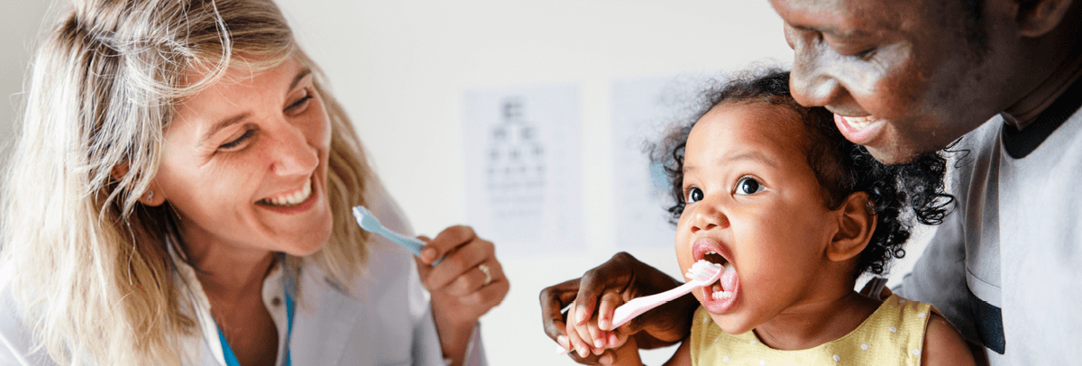female dentist showing father and baby daughter how to use toothbrush